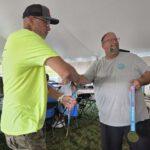 Two men shake hands under a tent at an awards event, one in a neon yellow shirt and the other holding a medal with a purple ribbon in hand.