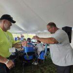 Two men fist-bump under a large white tent at an outdoor event, with seating, medals, and people in the background.