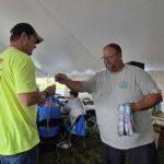 Two men exchange medals at an outdoor award event under a white tent, with trophies on a table in the background.