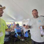 Two men under a white tent fist-bump, wearing lanyards and medals after a race event.