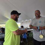 Two men under a white tent shaking hands; one in neon shirt, the other in gray with a large gold medal, smiling.