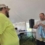Bearded man in a gray shirt shows gold medals on blue ribbons to another person inside a white tent event hall.
