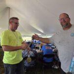 Two men under a large white tent at an outdoor event; one in a neon yellow shirt with a medal around his neck, the other in a gray shirt handing him something.