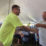 Two men shake hands under a large white tent at an outdoor event; one wears a bright lime shirt and sunglasses, smiling at the other.