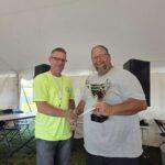 Two men shake hands under a white tent; one in a neon shirt presents a silver trophy to the other in a gray shirt, smile on both faces.