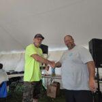 Two men shake hands under a white tent; one in a neon shirt presents a trophy at an outdoor event.