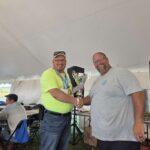 Two men shake hands and share a trophy at an outdoor awards moment under a white tent, smiling for the camera.