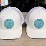 Two white trucker hats with teal circular patches on a wooden table, viewed from the front.