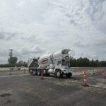 White concrete mixer truck navigating a cone-lined course in a large parking lot under a cloudy sky.