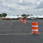 Row of white event tents set up in a large parking lot, with orange traffic barrels guiding a lane distance in front of them.