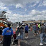 Crowd of people line up in a mall parking lot beside a large orange cement mixer truck during an outdoor event on a sunny day.