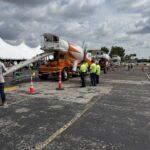 Line of cement mixer trucks with chutes extended at an outdoor demonstration; workers in neon shirts stand near cones and tents under a cloudy sky.