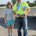 Man in a neon-green shirt with a trophy and medals stands beside a smiling woman in a gray t-shirt and teal shorts outdoors on a sunny day in a parking loted area.