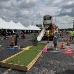Outdoor event with white tents, a wooden mini-golf hole, and an orange truck slide; people nearby.