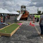 Portable mini-golf setup at an outdoor event, with a green padding lane on asphalt and an orange cement mixer with a slide behind it; tents and people in the background.