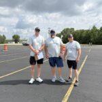 Three volunteers wearing gray T-shirts and lanyards stand in a nearly empty parking lot with orange traffic cones in the background.