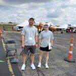 Two volunteers in gray shirts and lanyards pose on an airfield with white tents and aircraft in the background.