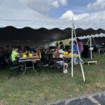 People gathered under a large white canopy for an outdoor event, sitting at folding tables with yellow helmets and food nearby on the tables.