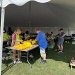 Children work on yellow plastic bins at a craft table under a large white event tent outdoors.