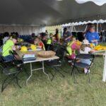 Children and adults craft at folding tables under a white canopy, with yellow safety vests and hard hats nearby on the grass campsite.