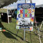 Large colorful sign reading "KID'S CORNER" under a white event tent at a truck championship event stage area in the grass.
