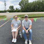 Two volunteers in gray shirts sit on folding chairs in a parking lot, with colorful pennants strung behind them.
