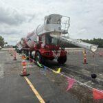Red and white concrete mixer truck with an extended chute in a paved lot, surrounded by orange cones and colorful pennant tape.