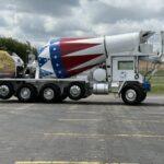 White cement mixer truck with a large patriotic red‑white‑blue drum; trailer in back carrying a banner reading “We the People.”