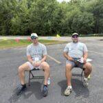 Two men sit on folding chairs in a parking lot, wearing gray shirts and caps with lanyards, holding cups while awaiting event.