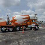 Orange and white concrete mixer truck on a flatbed trailer in a parking lot, with a person inspecting the discharge chute and traffic cones nearby.