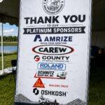 Large sponsor board at an outdoor event reads 'THANK YOU TO OUR PLATINUM SPONSORS' with logos for Amrize, Carew, County Materials, Roland Machinery Co., Schmitz Ready Mix, Sika, and Oshkosh.