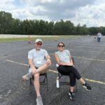 Two volunteers sit on folding chairs in a large parking lot with orange cones and trees in the background.
